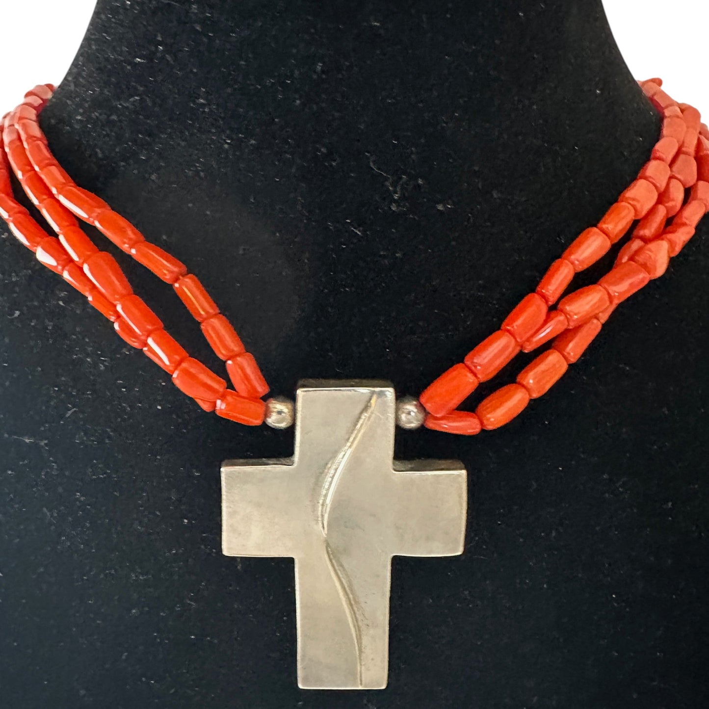 Close-up of flat sterling silver cross pendant with engraved sinuous line detail on face, suspended from coral bead strands